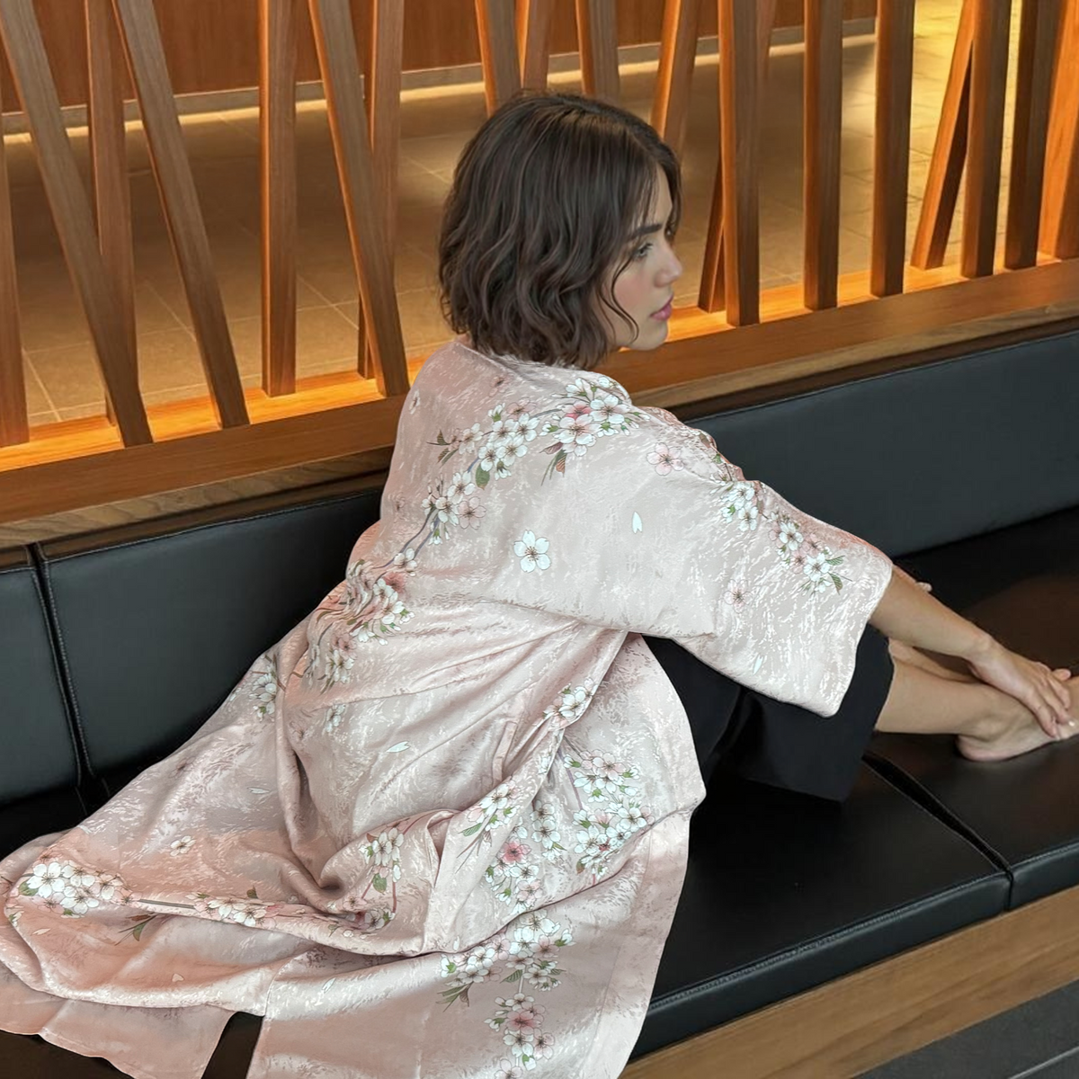 Sakura Pink silk kimono robe with cherry blossom motif, worn seated on a dark banquette against a warm wood-slat wall